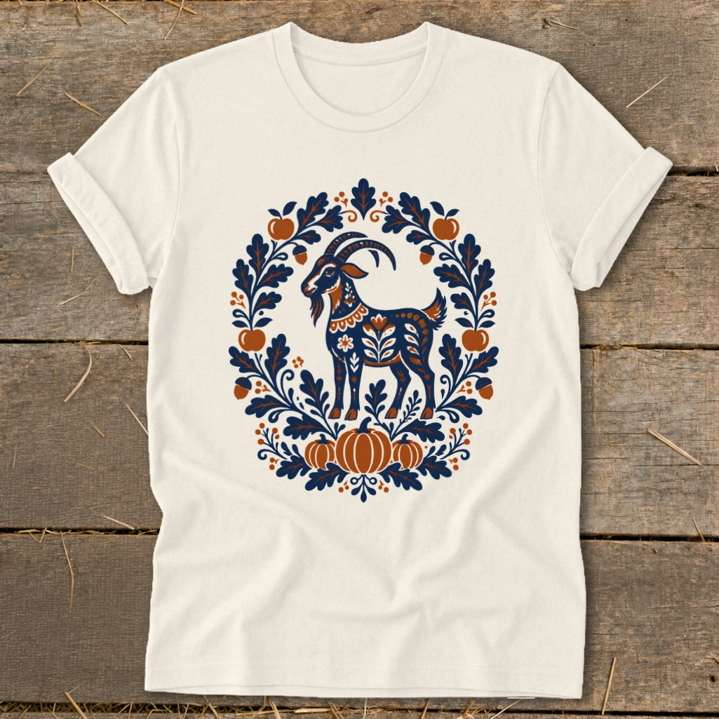 A cream t-shirt features a folk-art goat inside an autumn-themed wreath of leaves, acorns, and pumpkins.