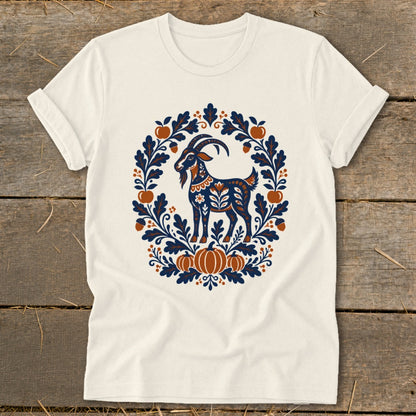A cream t-shirt features a folk-art goat inside an autumn-themed wreath of leaves, acorns, and pumpkins.