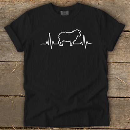 A black crewneck t-shirt featuring a white graphic of a sheep silhouetted against a heartbeat line, displayed flat on a wooden surface.