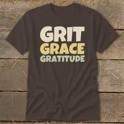 A dark brown t-shirt featuring the stacked text Grit, Grace, and Gratitude in cream, yellow, and green.