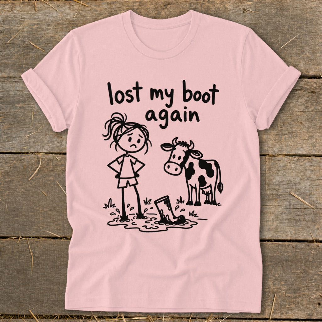 A pink t-shirt featuring a cartoon of a sad girl and a cow standing near a muddy boot, with the text "lost my boot again."