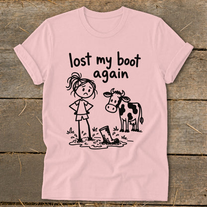 A pink t-shirt featuring a cartoon of a sad girl and a cow standing near a muddy boot, with the text "lost my boot again."