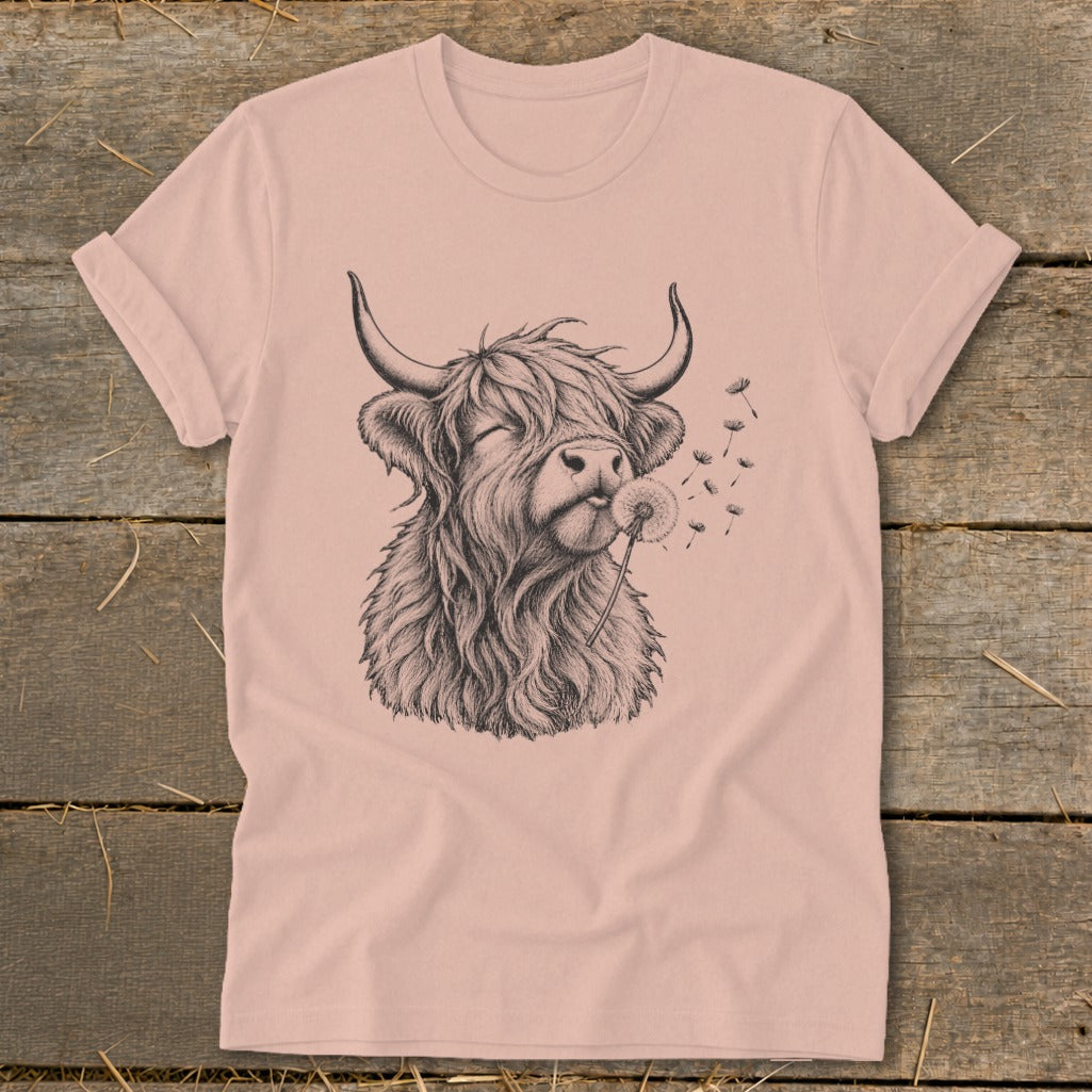 Highland cow with eyes closed blowing dandelion seeds on a light pink t-shirt.