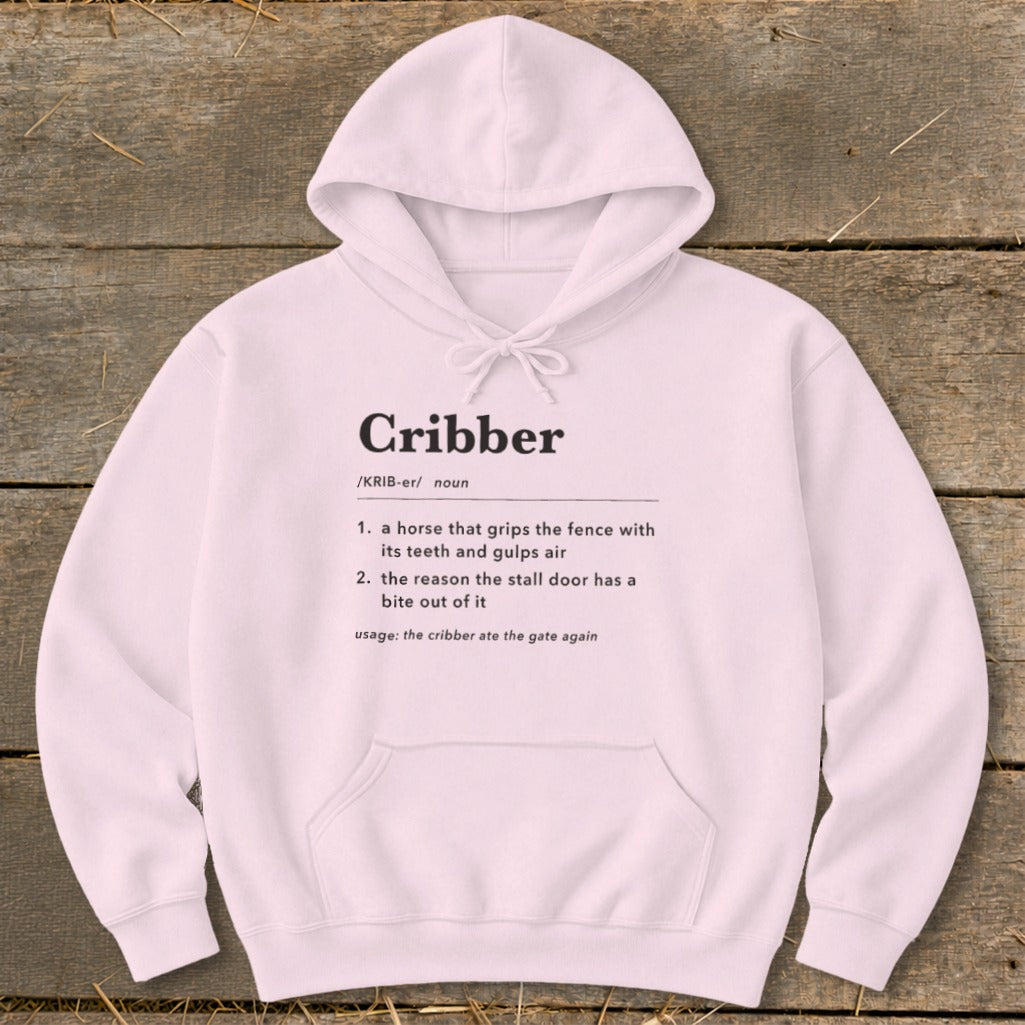 A light pink hooded sweatshirt with black text defining the word "Cribber" on the front, laid flat on a wooden background.