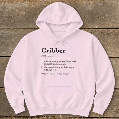 A light pink hooded sweatshirt with black text defining the word "Cribber" on the front, laid flat on a wooden background.