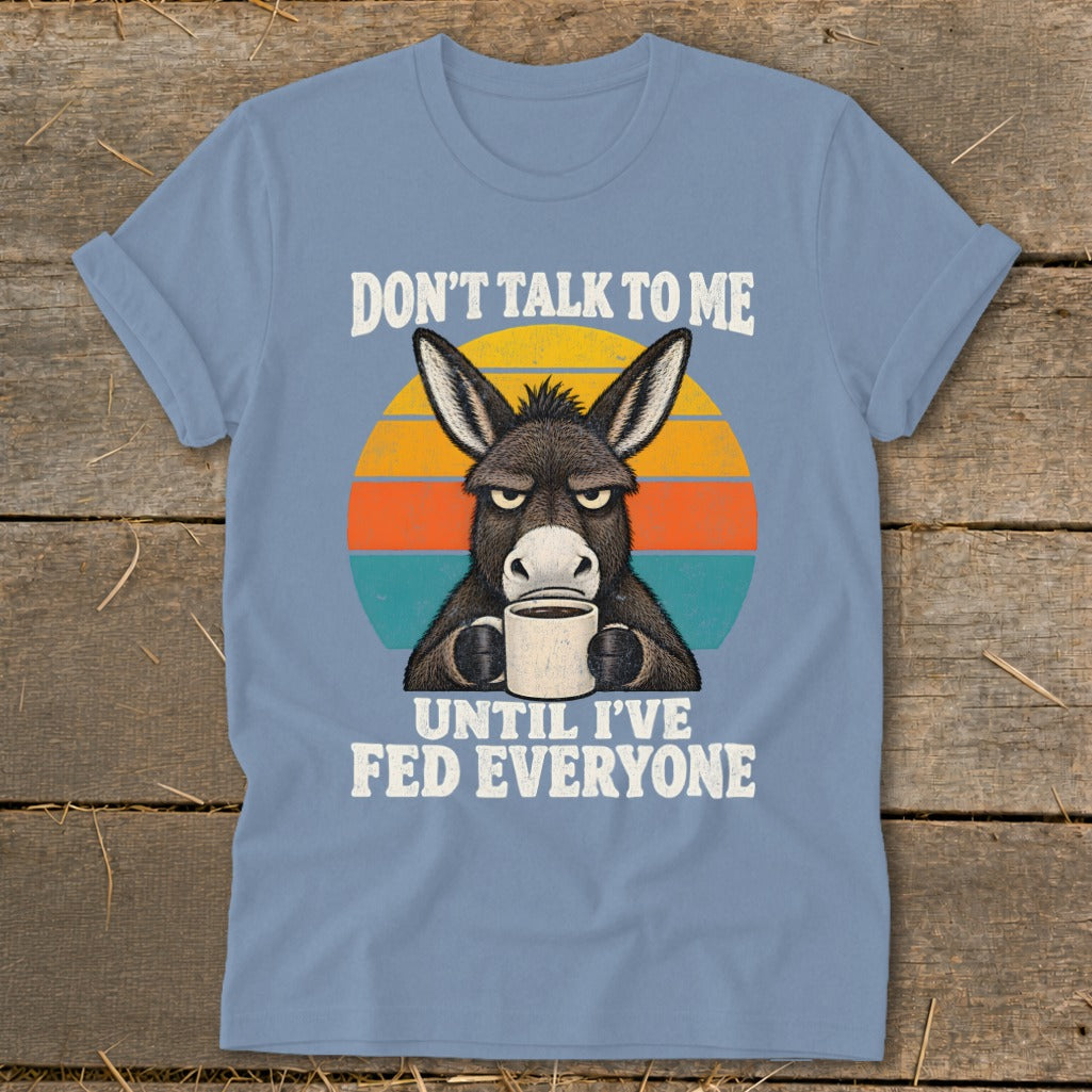 A light blue t-shirt featuring a grumpy donkey drinking coffee and text reading Don't Talk To Me Until I've Fed Everyone.