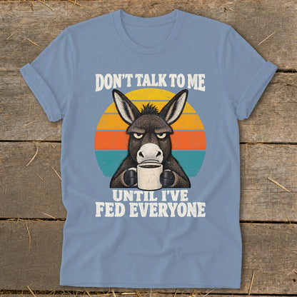 A light blue t-shirt featuring a grumpy donkey drinking coffee and text reading Don't Talk To Me Until I've Fed Everyone.