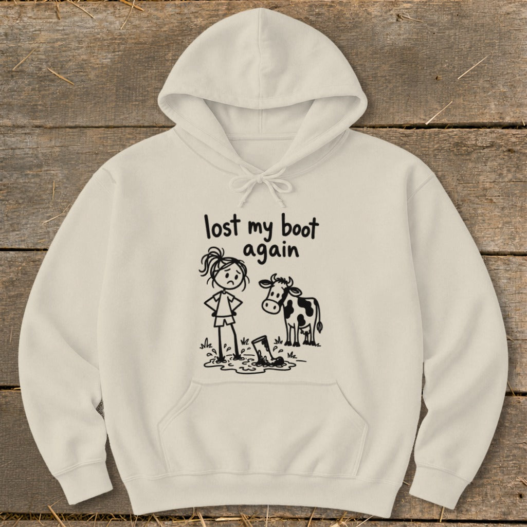 Cream-colored hooded sweatshirt featuring a cartoon graphic of a sad girl and cow next to a boot stuck in a puddle, with the text lost my boot again.