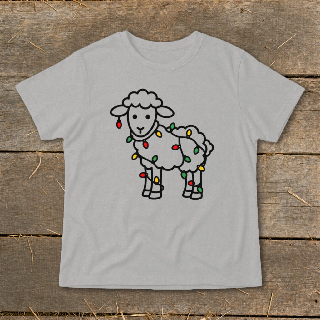 A light gray t-shirt featuring a black outline drawing of a fluffy sheep wrapped in colorful Christmas lights, laid flat on a rustic wooden surface.