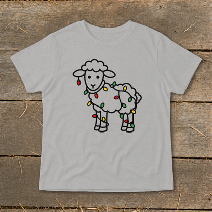 A light gray t-shirt featuring a black outline drawing of a fluffy sheep wrapped in colorful Christmas lights, laid flat on a rustic wooden surface.