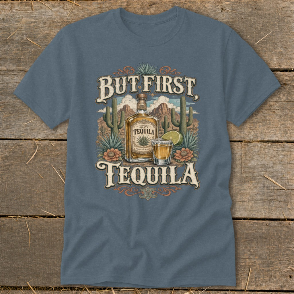 Blue t-shirt graphic with tequila bottle, shot glass, cacti, and desert landscape. Text reads "But First, Tequila."