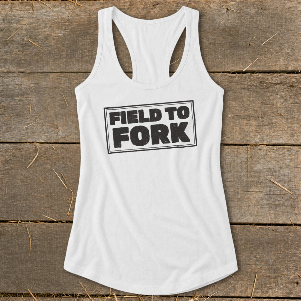 White racerback tank top featuring the black text Field to Fork inside a bordered rectangle, laid on a rustic wooden background.