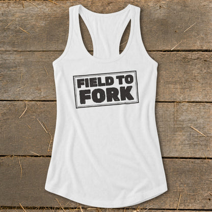 White racerback tank top featuring the black text Field to Fork inside a bordered rectangle, laid on a rustic wooden background.