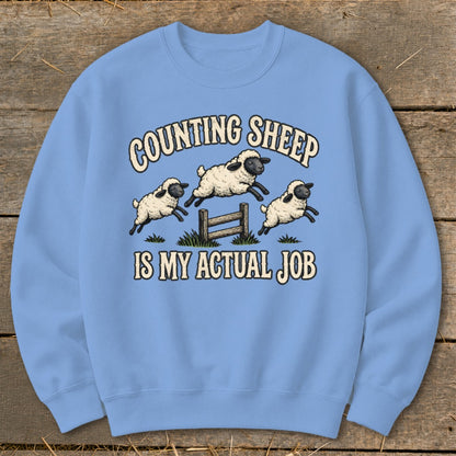 Light blue sweatshirt with illustration of sheep jumping over a fence and text "COUNTING SHEEP IS MY ACTUAL JOB".