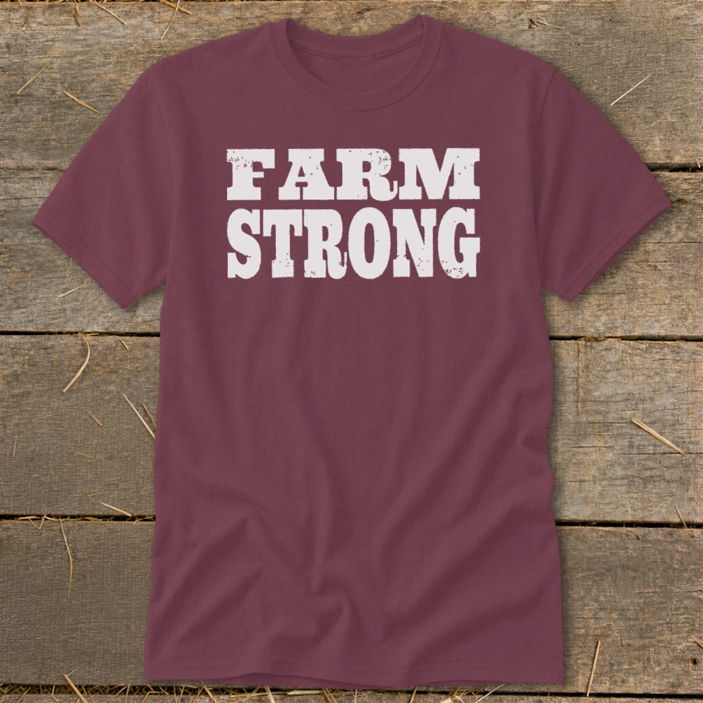 Maroon crewneck t-shirt featuring the distressed white text FARM STRONG across the chest, laid flat on a weathered wooden surface.