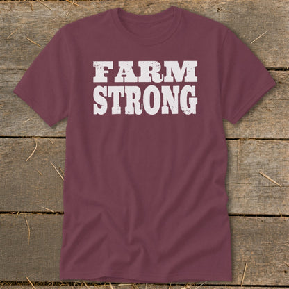 Maroon crewneck t-shirt featuring the distressed white text FARM STRONG across the chest, laid flat on a weathered wooden surface.