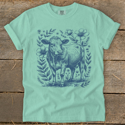 Teal t-shirt with a navy blue drawing of a cow surrounded by flowers and leaves.
