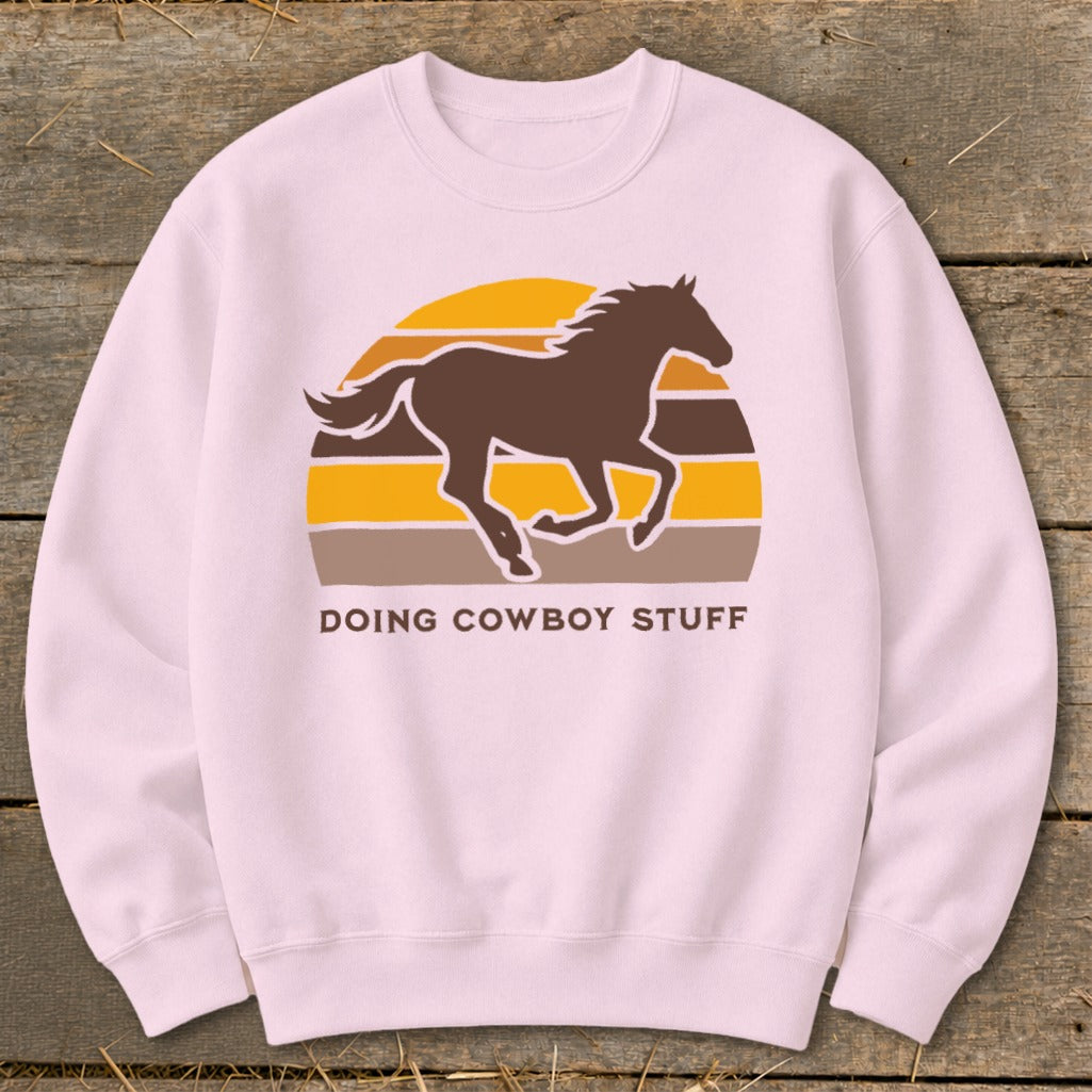 Pink sweatshirt with horse and sunset graphic, text reads "DOING COWBOY STUFF".