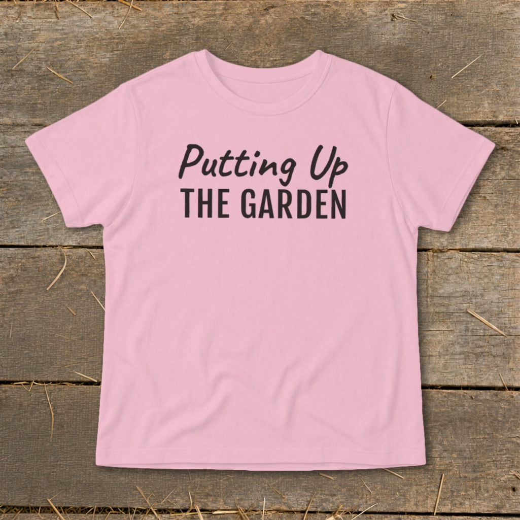 Pink t-shirt with black text "Putting Up THE GARDEN" on a wooden background.