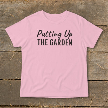 Pink t-shirt with black text "Putting Up THE GARDEN" on a wooden background.