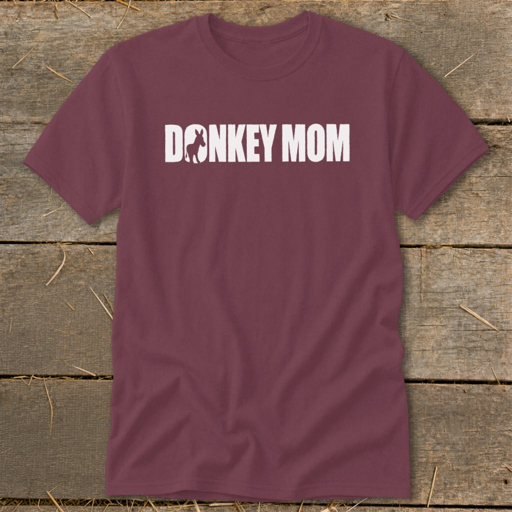 Maroon t-shirt with white text reading "Donkey Mom," featuring a donkey silhouette inside the letter O, laid on wooden planks.
