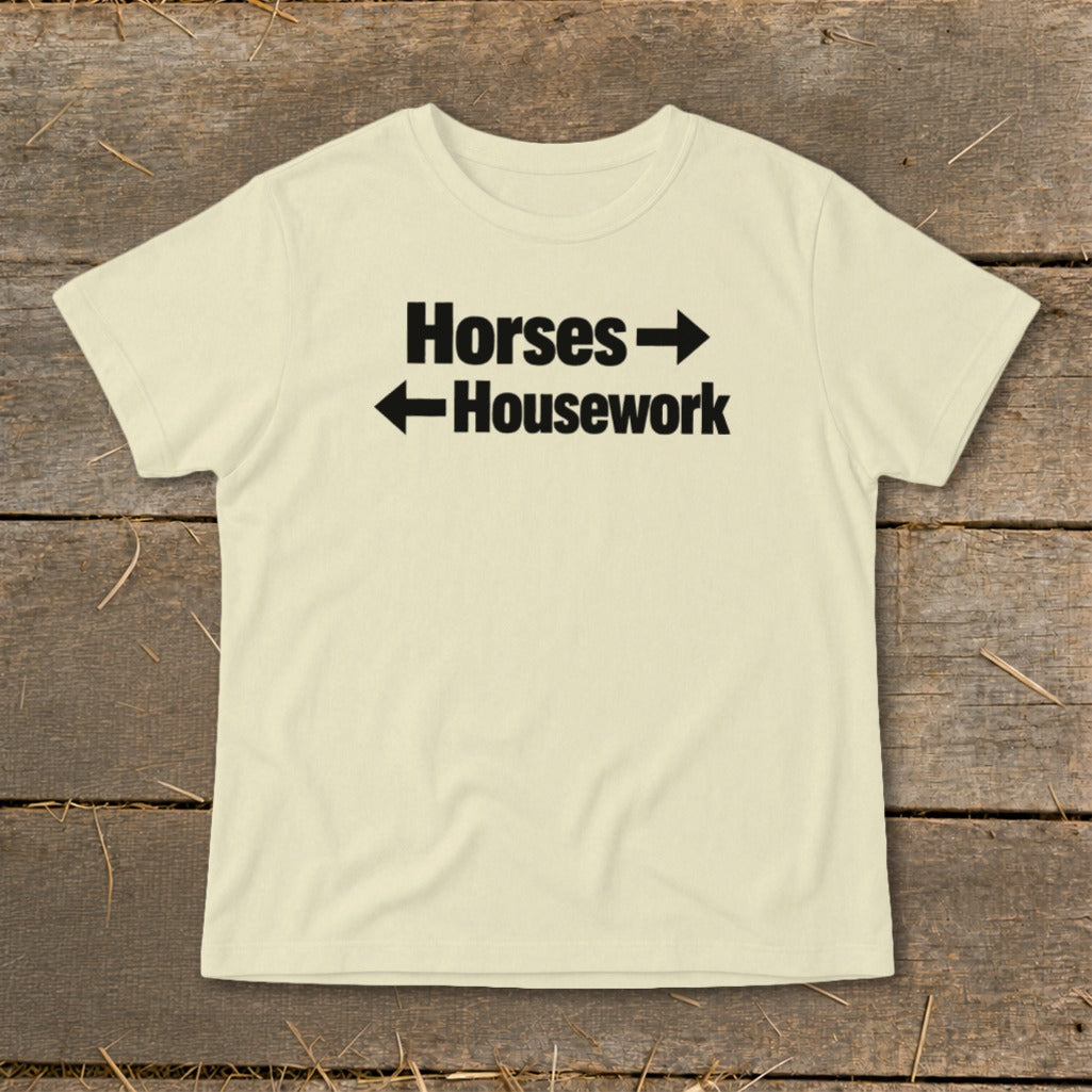 A cream-colored t-shirt with black text reading Horses with a right arrow and Housework with a left arrow, laid flat on a rustic wooden surface.