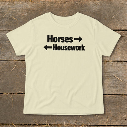 A cream-colored t-shirt with black text reading Horses with a right arrow and Housework with a left arrow, laid flat on a rustic wooden surface.