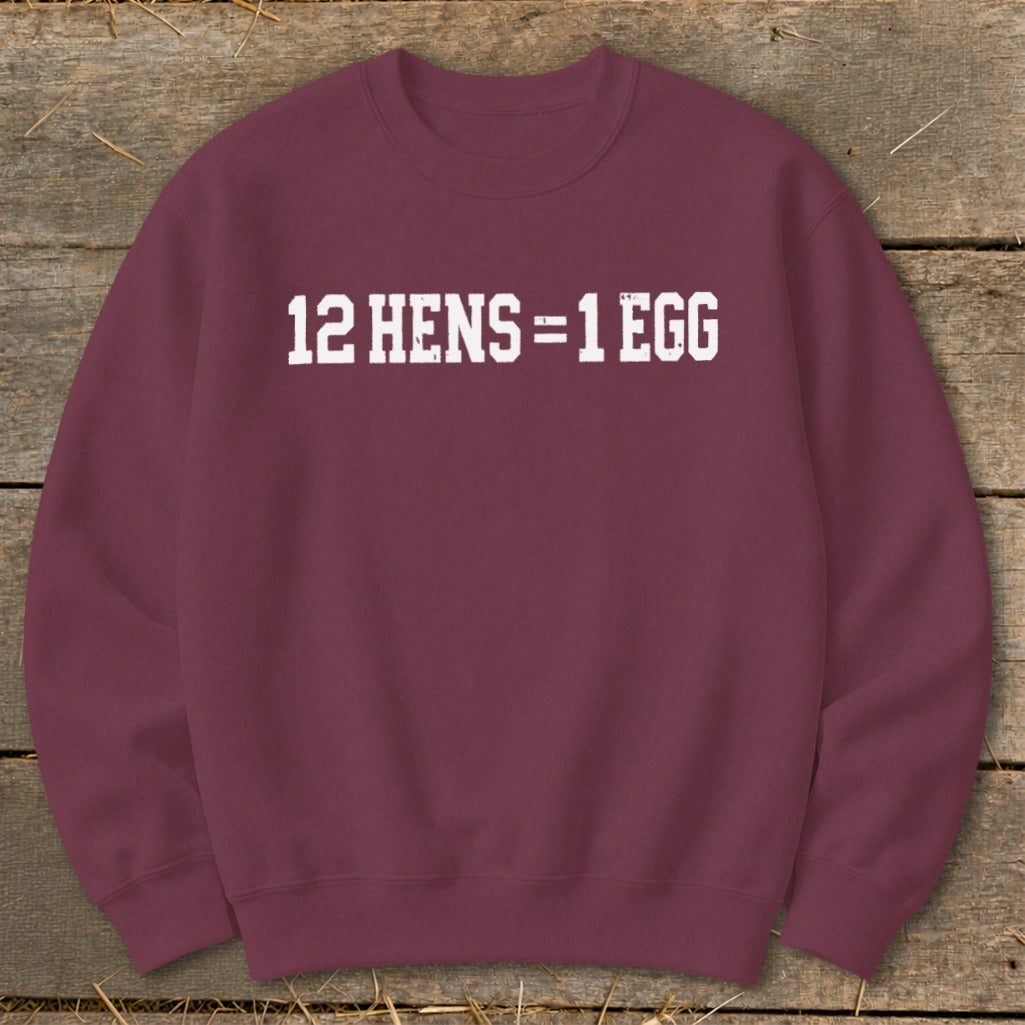 Maroon crewneck sweatshirt featuring distressed white text that reads 12 HENS = 1 EGG, laid flat on a rustic wooden surface.
