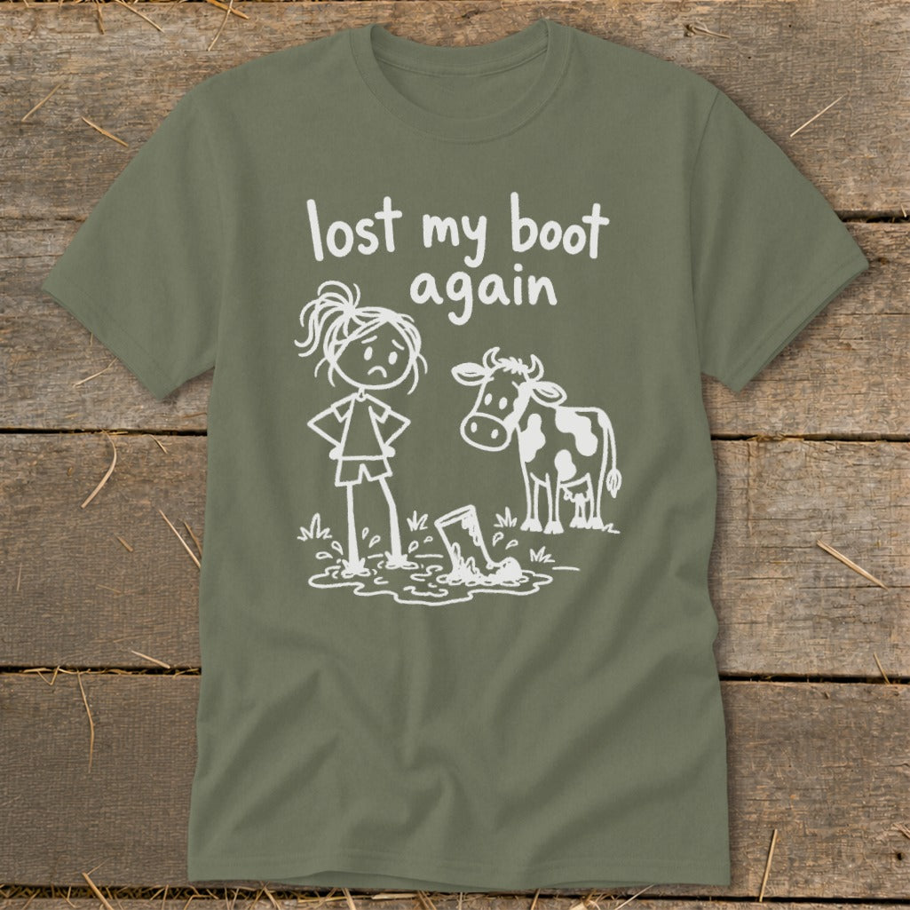 An olive green t-shirt featuring a white stick figure graphic of a girl and a cow standing in a mud puddle next to a lost boot, with the text "lost my boot again."
