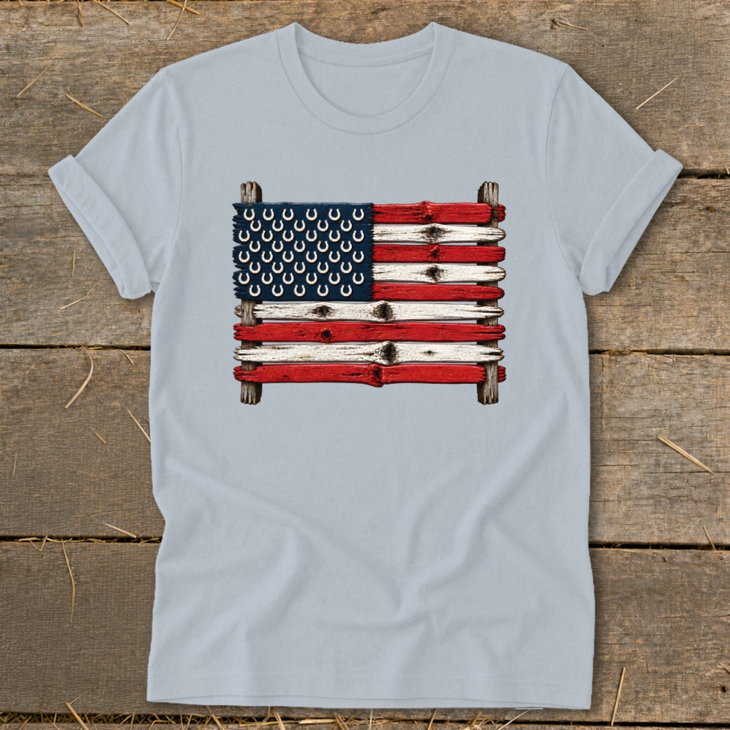 Light blue t-shirt featuring a graphic of the American flag made of painted wooden planks and horseshoes, laid on a rustic wooden background.