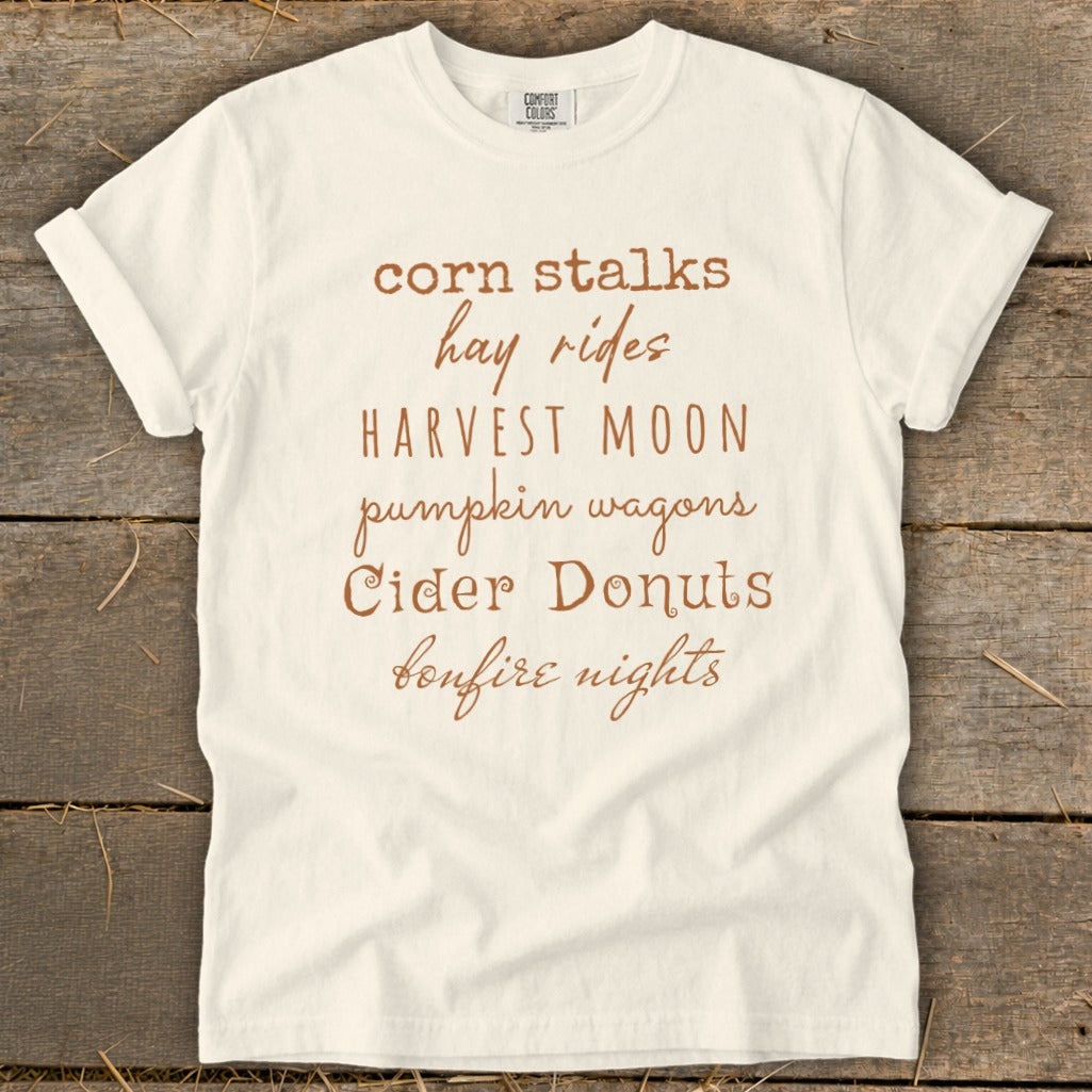 Cream t-shirt with brown text listing fall activities: corn stalks, hay rides, harvest moon, pumpkin wagons, cider donuts, bonfire nights.